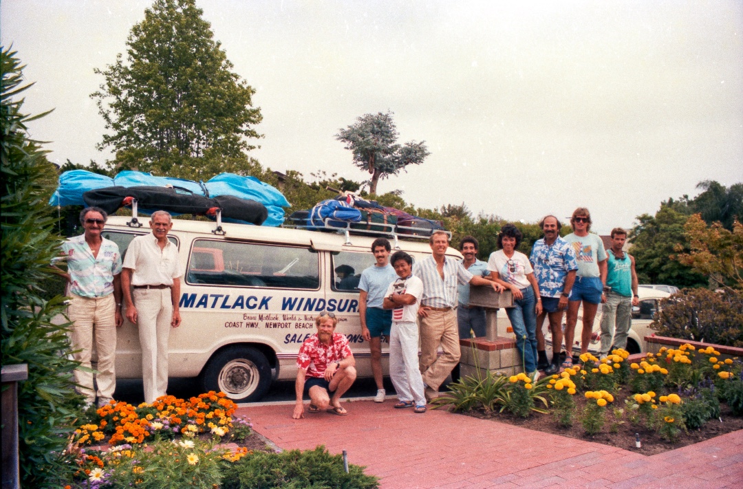 Early 1980s photo of Bruce Matlack, crouched down in a red shirt, alongside his Matlack Windsurfing van and a group of enthusiastic tourists.