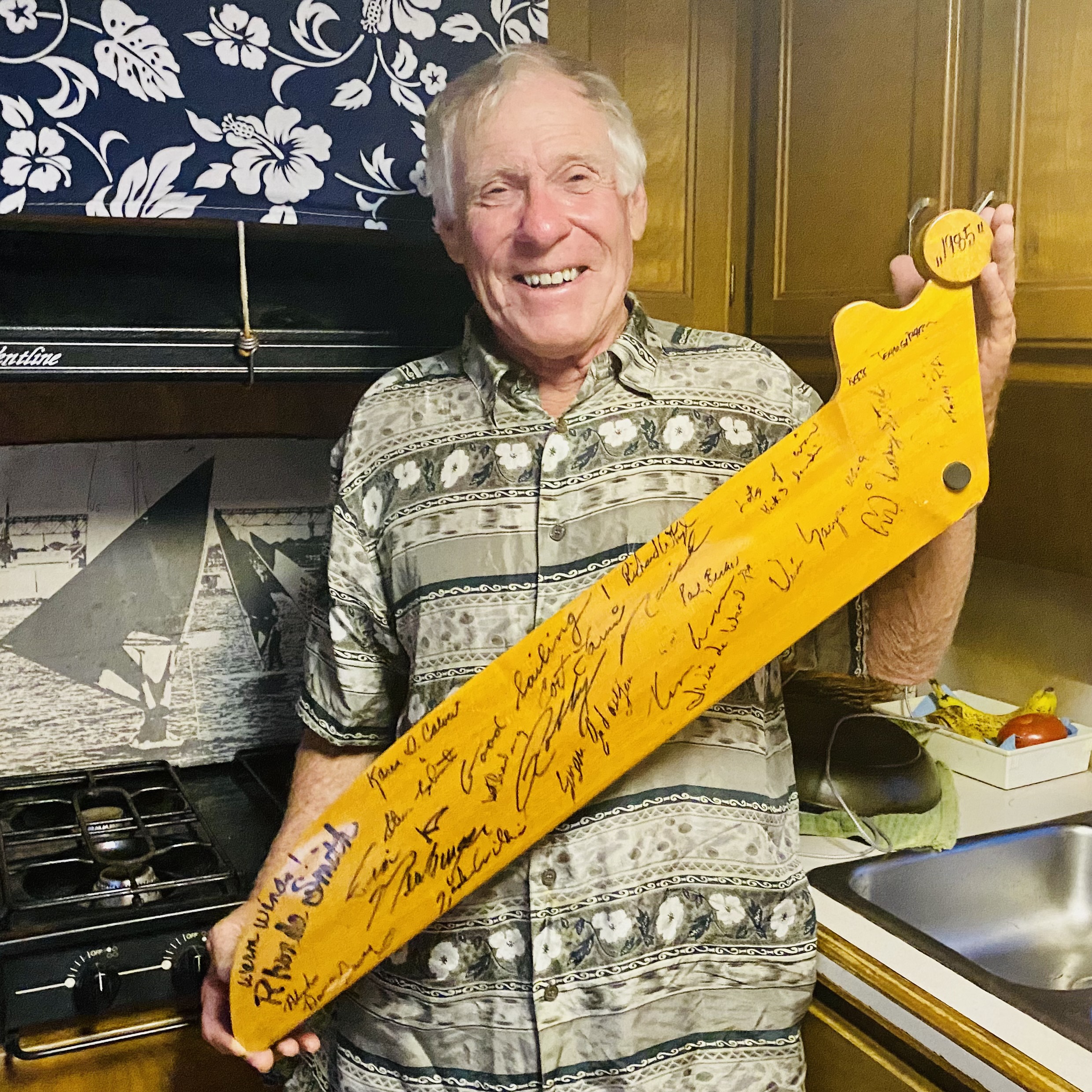 Bruce Matlack smiles as he holds a daggerboard signed by windsurfing luminaries of the 1980s, commemorating the community and competitive spirit that propelled the sport forward.