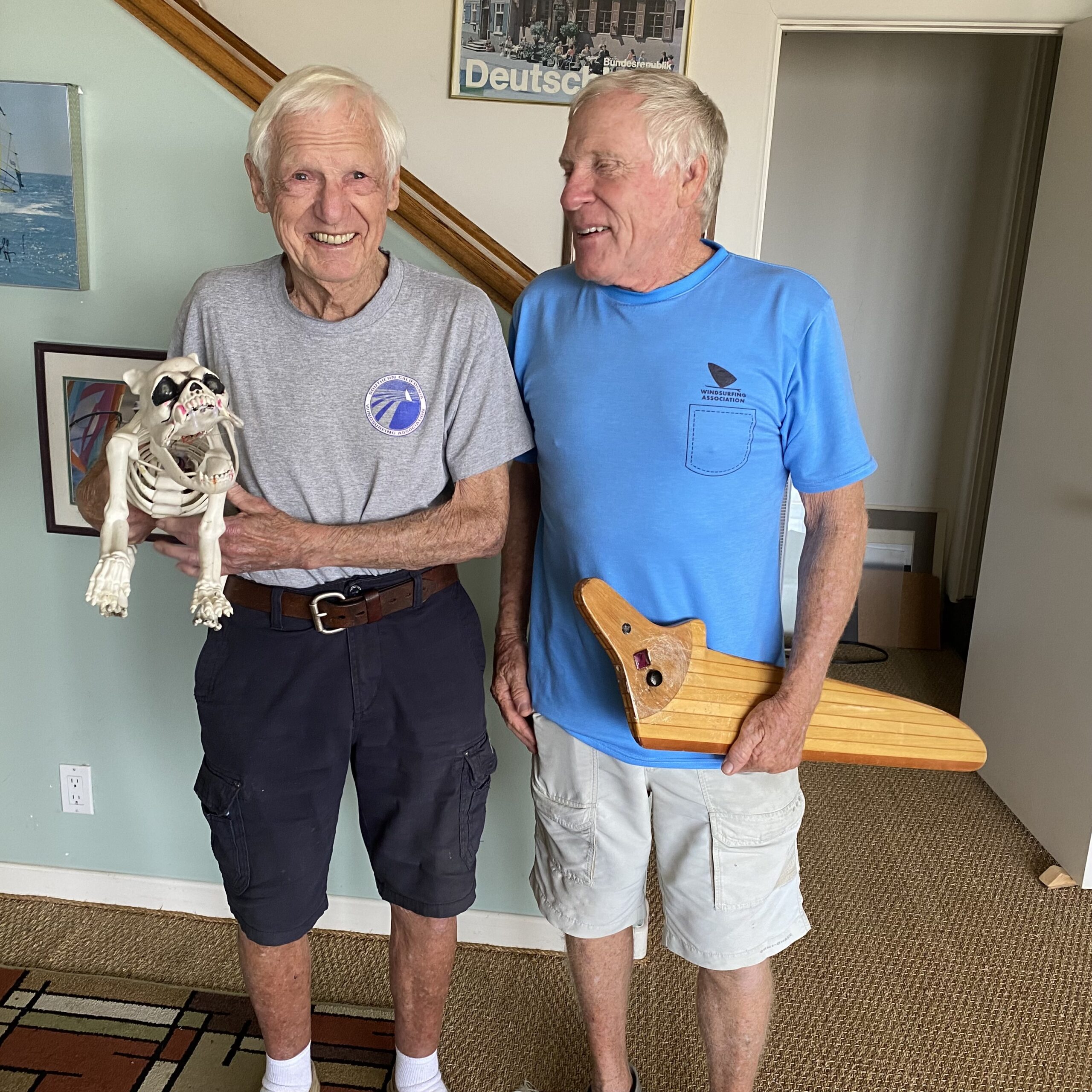 Bruce Matlack and Paul Hengstebeck standing together in Laguna Beach, 2021, holding a vintage windsurfing board and a skeleton model, celebrating the legacy of windsurfing.